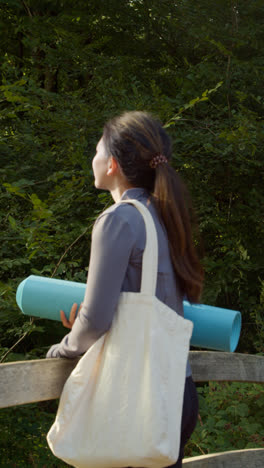 Vertical-Video-Of-Woman-Wearing-Sports-Clothing-Carrying-Yoga-Mat-Standing-On-Bridge-In-Forest-Looking-Around-Enjoying-Peace-And-Beauty-Of-Nature-Shot-In-Real-Time